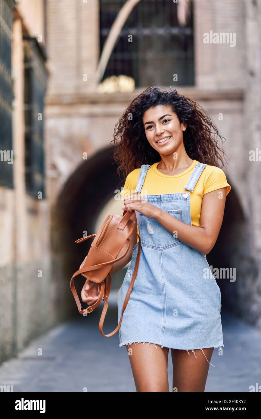 Giovane turista araba con capelli ricci all'aperto. Arab viaggiatore ragazza in abiti casual per la strada. Donna felice con t-shirt gialla e abito in denim su sfondo urbano. Giovane donna turistica nera con capelli ricci all'aperto Foto Stock