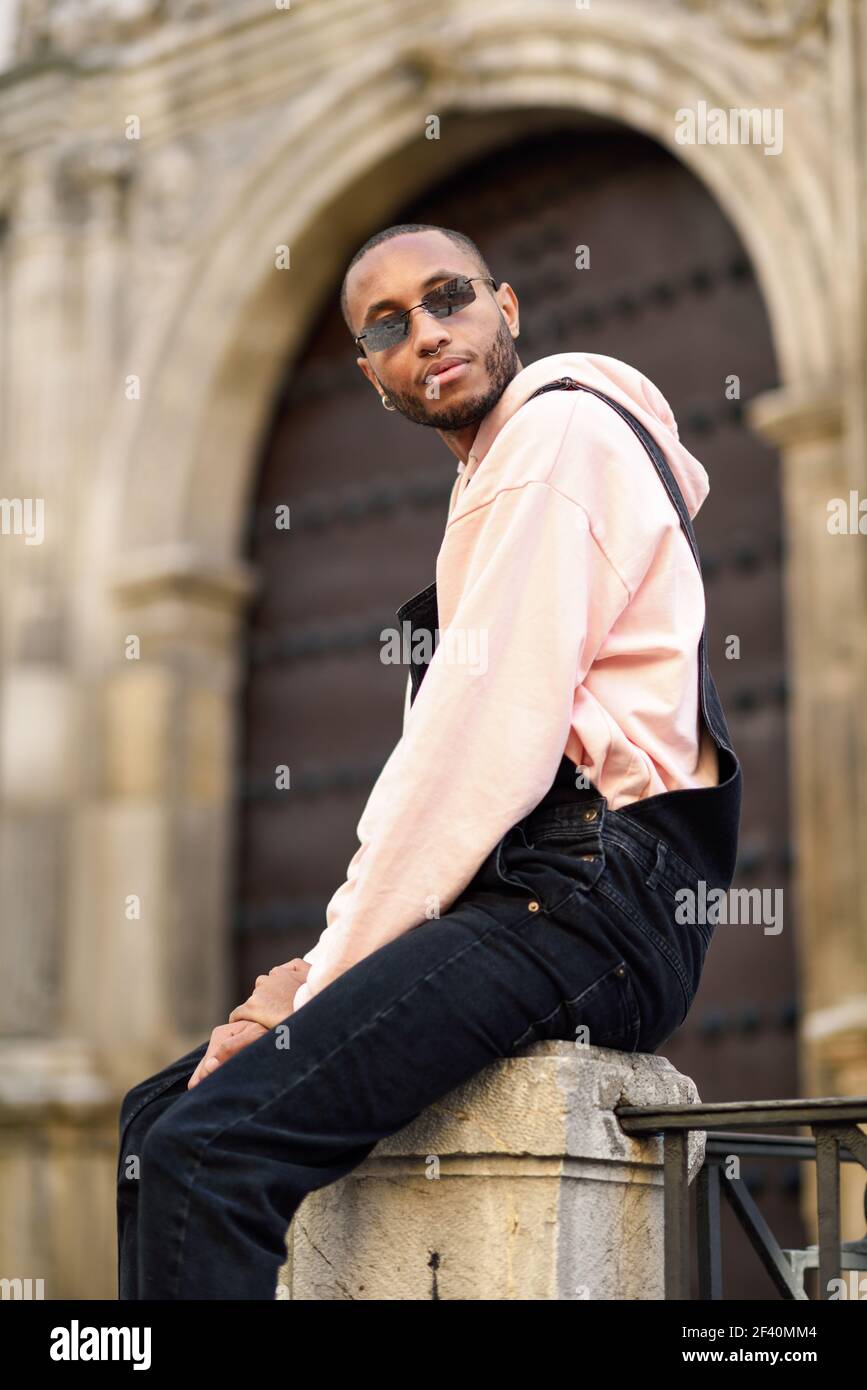 Giovane uomo nero che indossa abiti casual e occhiali da sole seduti sulla strada. Ragazzo africano con pantaloni di bib all'aperto. Giovane uomo nero che indossa abiti casual e occhiali da sole seduti sulla strada Foto Stock