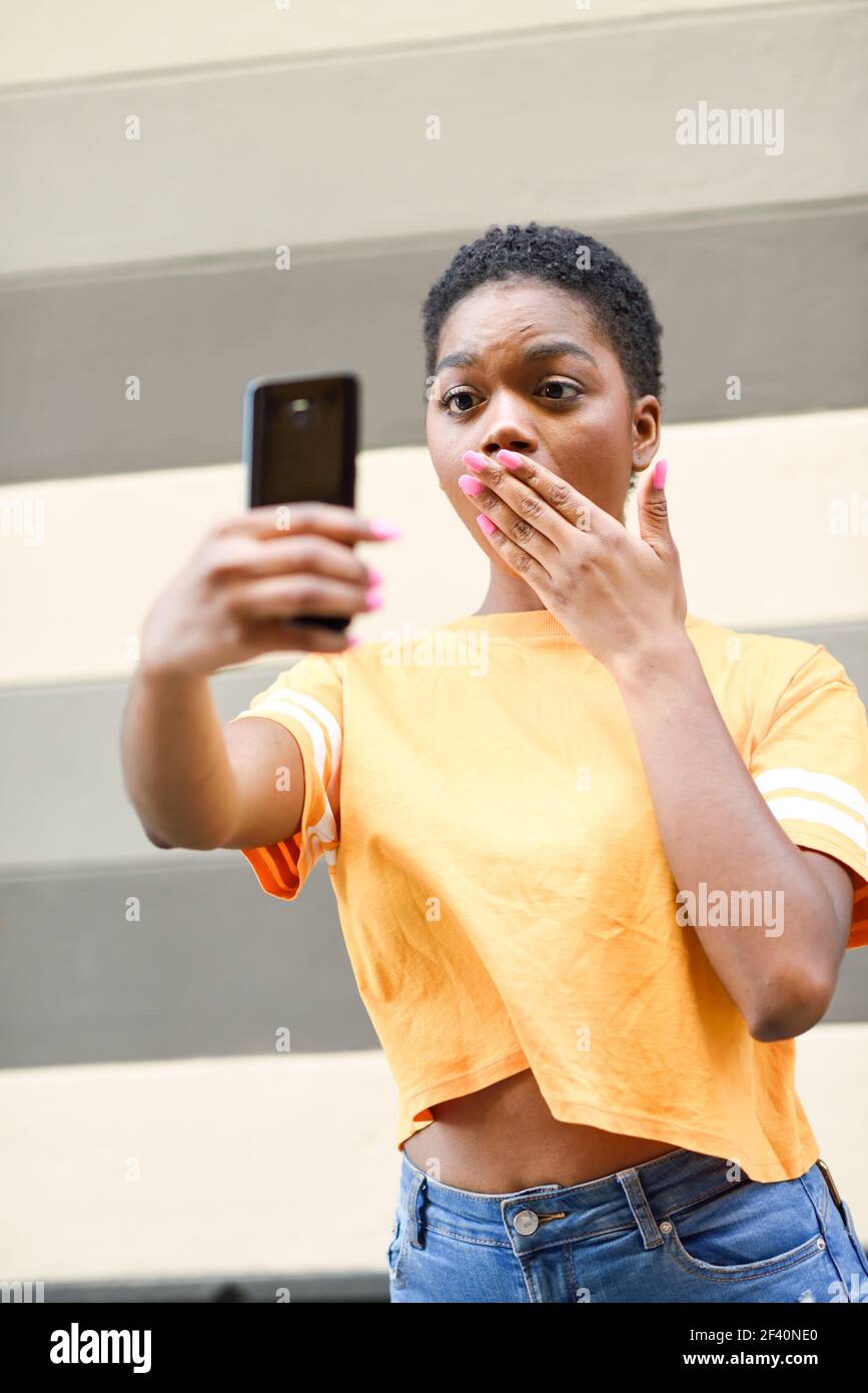 Giovane donna nera che scatta foto selfie con l'espressione divertente all'aperto. Ragazza con capelli molto corti che indossa abiti casual.. Giovane donna nera che scatta foto selfie con l'espressione divertente all'aperto. Foto Stock