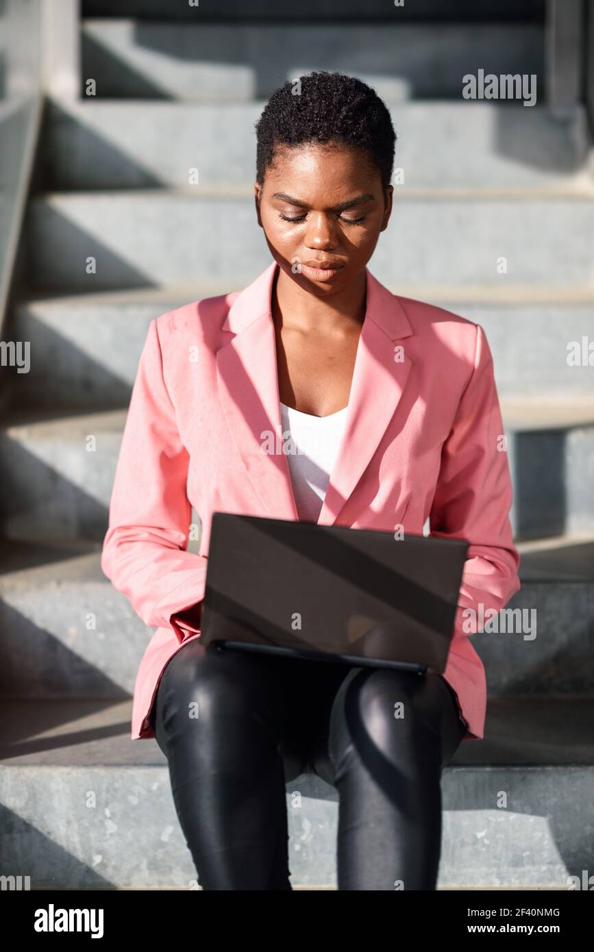 Donna nera seduta su gradini urbani che lavora con un computer portatile. Donna d'affari afroamericana che indossa un vestito con giacca rosa. Donna nera seduta su gradini urbani che lavora con un computer portatile. Foto Stock