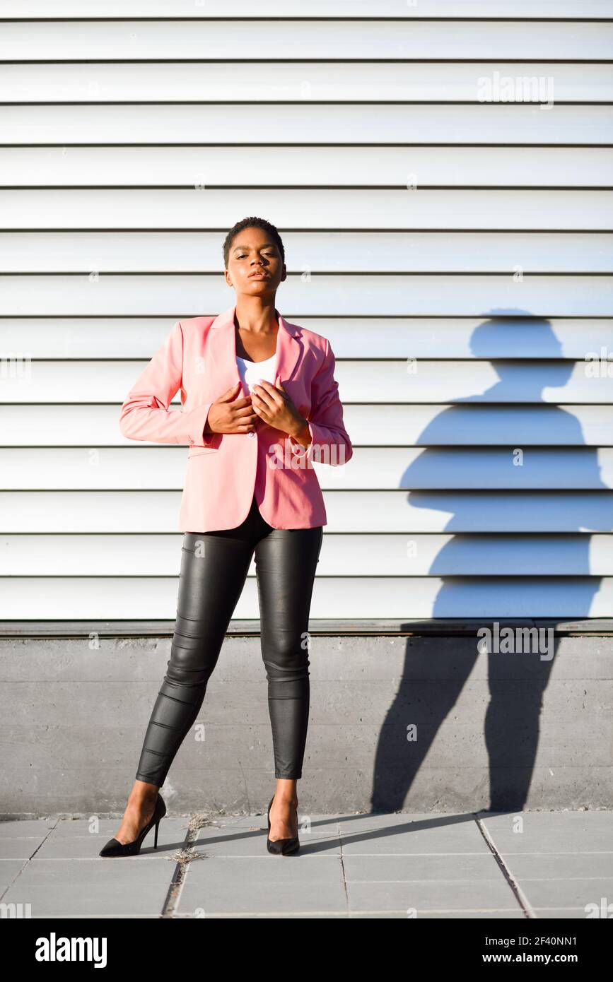 Donna nera, modello di moda, in piedi sulla parete urbana. Donna afroamericana con giacca rosa con luce al tramonto. Donna nera, modello di moda, in piedi sulla parete urbana Foto Stock