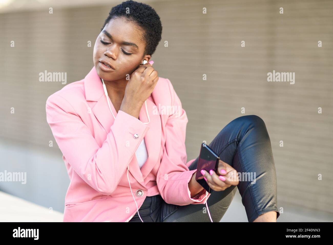 Donna d'affari nera seduta all'aperto utilizzando smartphone con auricolari. Donna afroamericana con giacca rosa. Donna d'affari nera seduta all'aperto utilizzando smartphone con auricolari Foto Stock