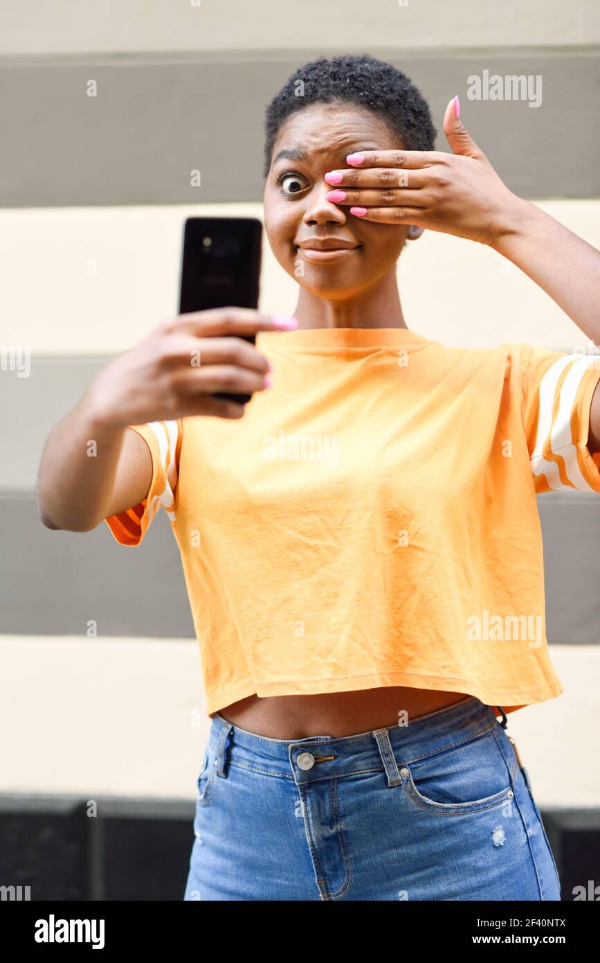 Divertente donna nera scattando foto selfie con felice espressione all'aperto. Ragazza con capelli molto corti che indossa abiti casual.. Divertente donna nera scattando foto selfie con felice espressione all'aperto Foto Stock