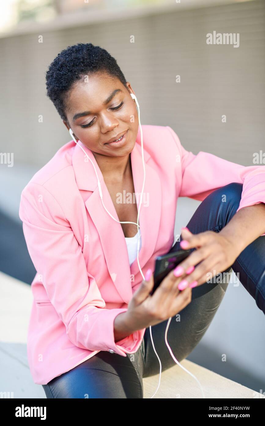 Donna d'affari nera seduta all'aperto che parla tramite videoconferenza con il suo smartphone. Donna afroamericana con giacca rosa. Donna d'affari nera seduta all'aperto che parla in videoconferenza con il suo smartphone. Foto Stock