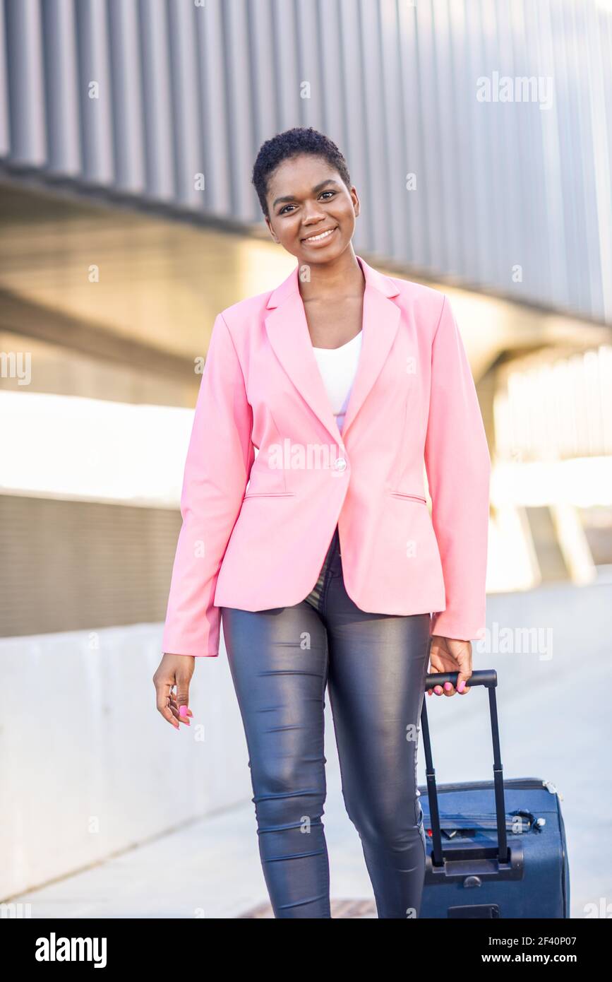 Donna d'affari afroamericana commuter con valigia a rulli vicino alla costruzione di ufficio di affari. Donna nera che cammina con borsa da viaggio che indossa giacca rosa. Donna nera che cammina con borsa da viaggio con giacca rosa. Foto Stock