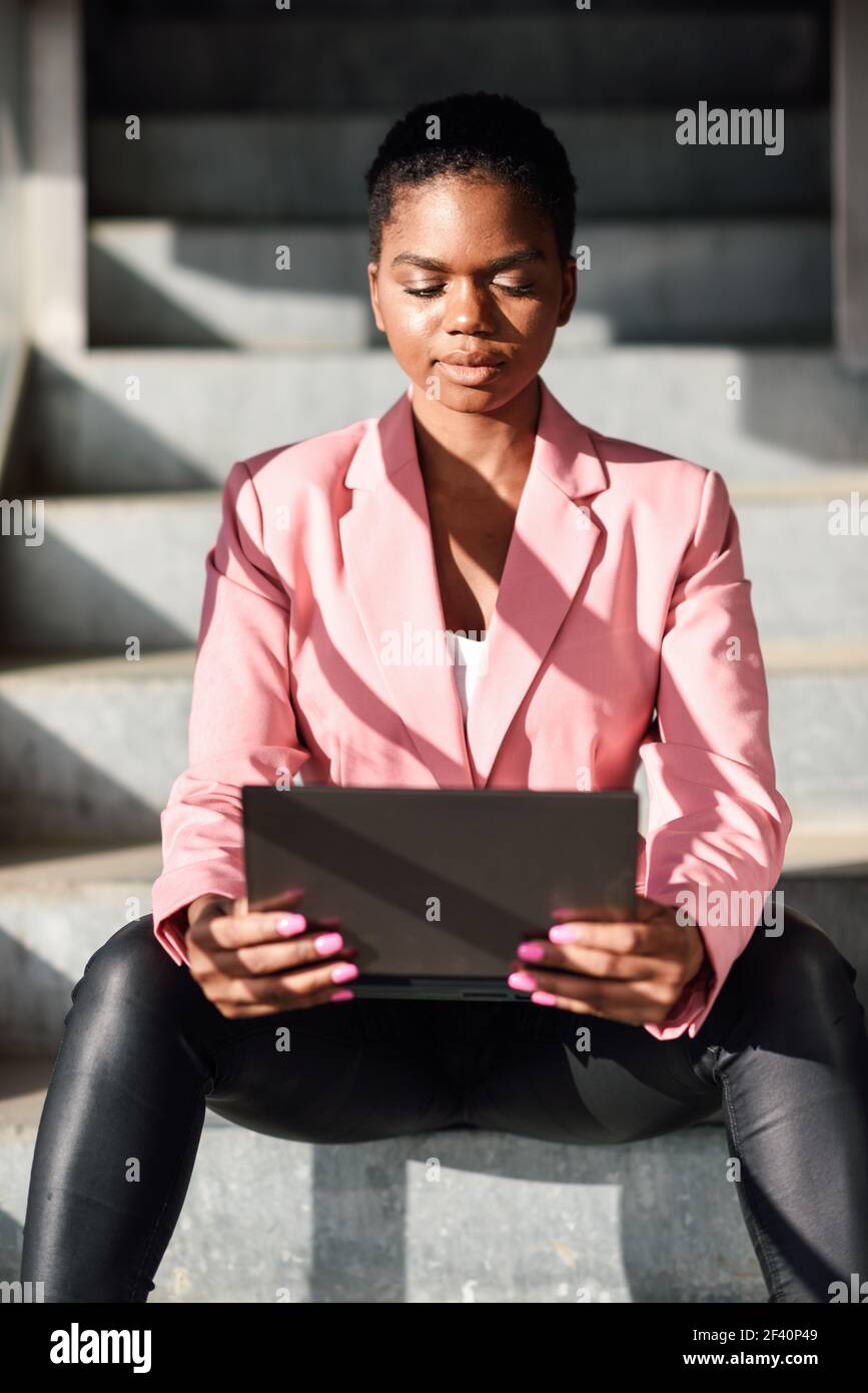 Donna d'affari nera seduta su gradini urbani lavorando con un computer portatile. Donna afroamericana con giacca rosa. Donna d'affari nera seduta su gradini urbani lavorando con un computer portatile. Foto Stock