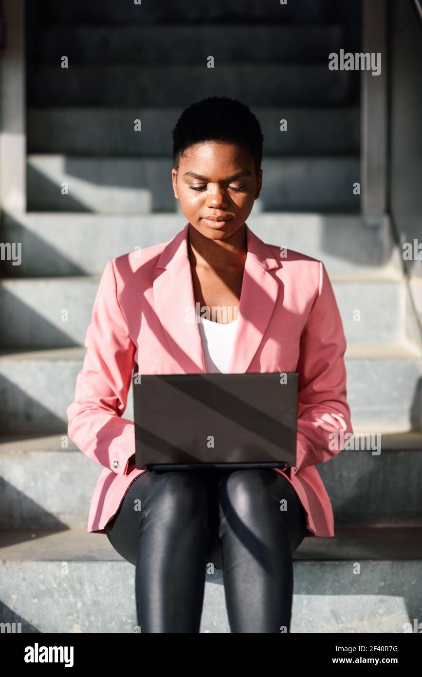 Donna nera seduta su gradini urbani che lavora con un computer portatile. Donna d'affari afroamericana che indossa un vestito con giacca rosa. Donna nera seduta su gradini urbani che lavora con un computer portatile. Foto Stock