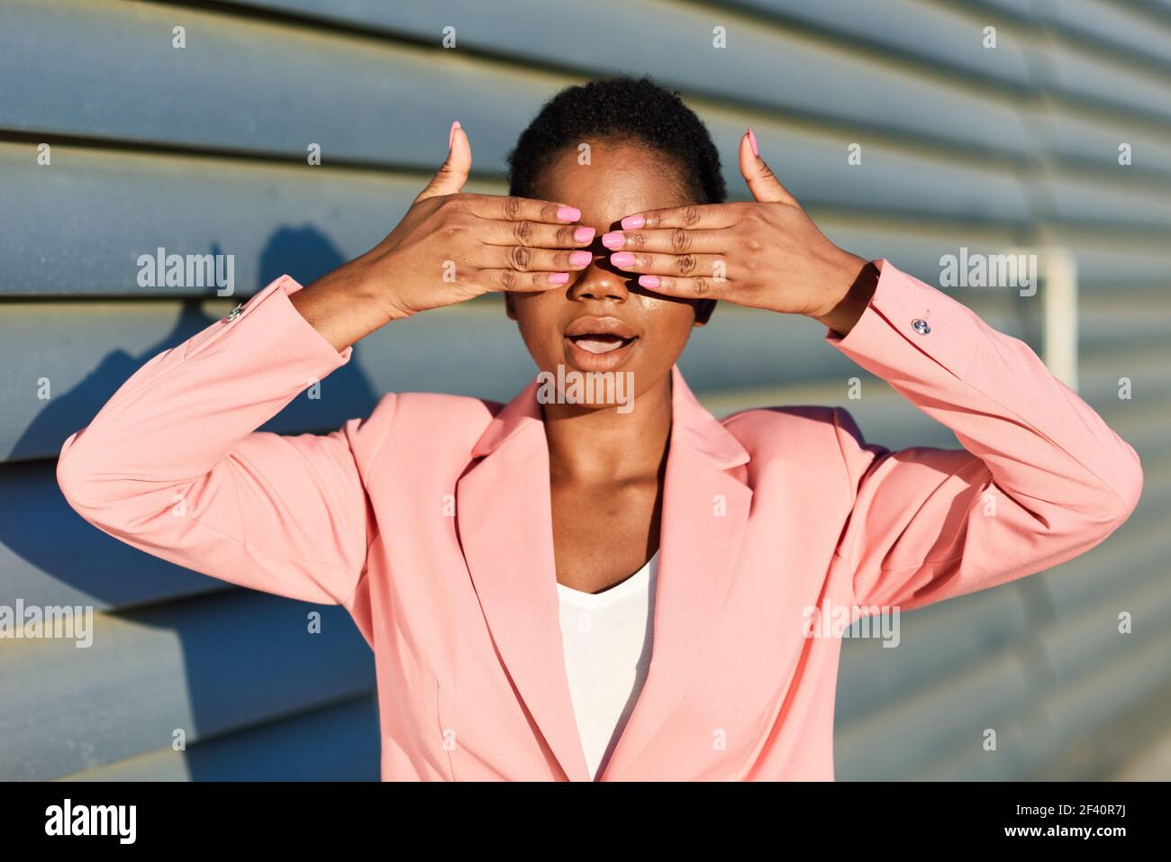 Donna nera divertente, modello di moda, in piedi sulla parete urbana che copre gli occhi con le mani. Donna afroamericana con giacca rosa con luce al tramonto. Donna nera divertente in piedi sulla parete urbana che copre i suoi occhi con le mani. Foto Stock
