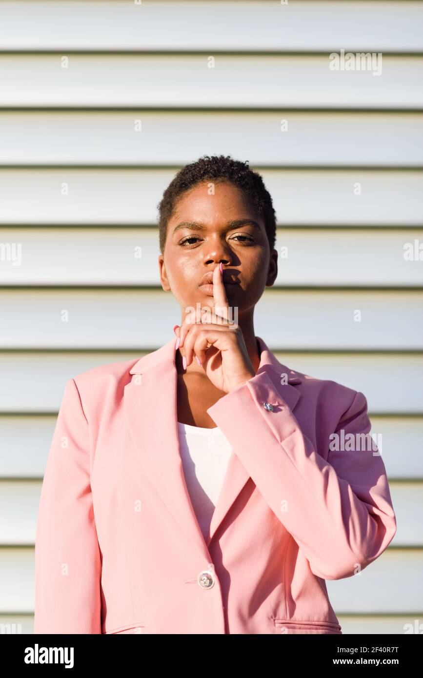 Donna d'affari nera che fa il gesto del silenzio. Donna afroamericana con giacca rosa con luce al tramonto. Donna d'affari afroamericana che fa il gesto del silenzio. Foto Stock