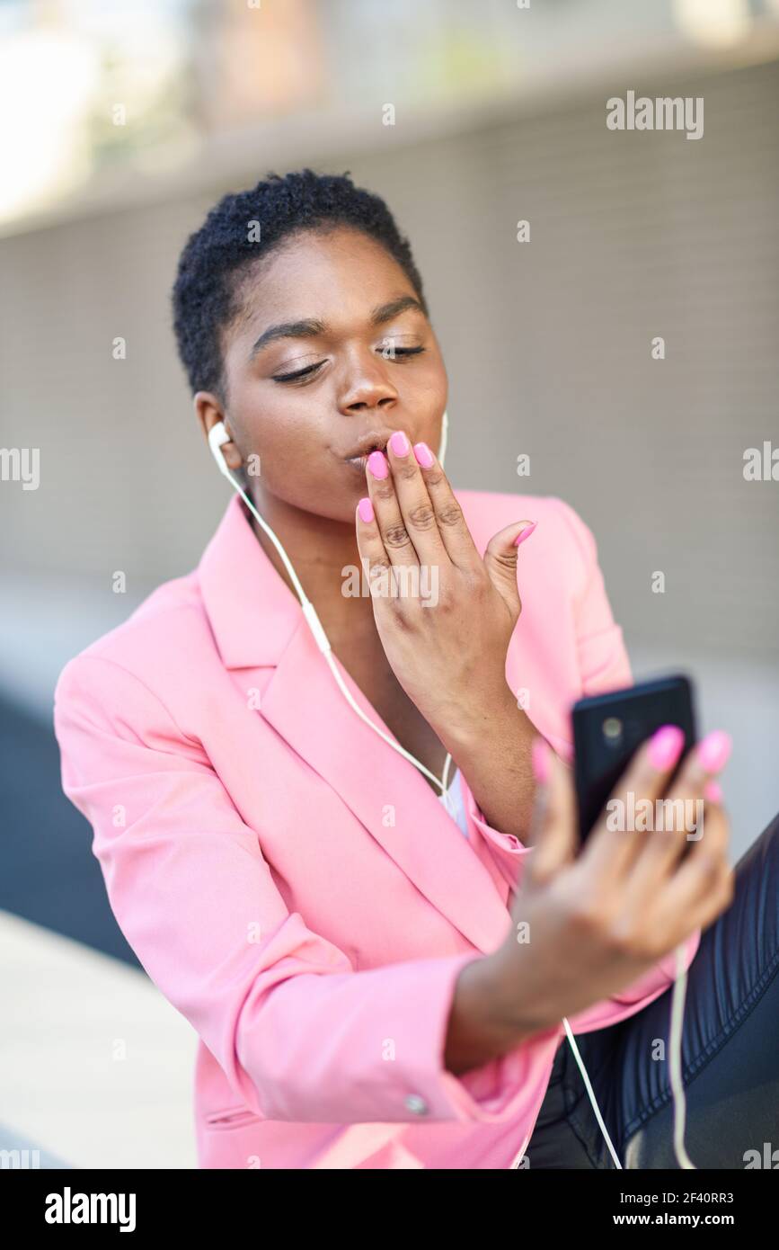 Donna d'affari nera seduta all'aperto che parla tramite videoconferenza con il suo smartphone. Donna afroamericana con giacca rosa. Donna d'affari nera seduta all'aperto che parla in videoconferenza con il suo smartphone. Foto Stock