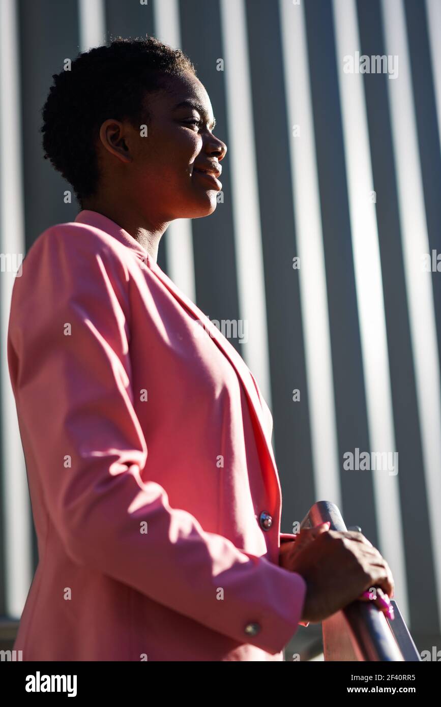 Donna d'affari nera sorridente in piedi vicino alla costruzione dell'ufficio di affari. Donna afroamericana con giacca rosa. Donna d'affari nera sorridente in piedi vicino alla costruzione dell'ufficio di affari Foto Stock