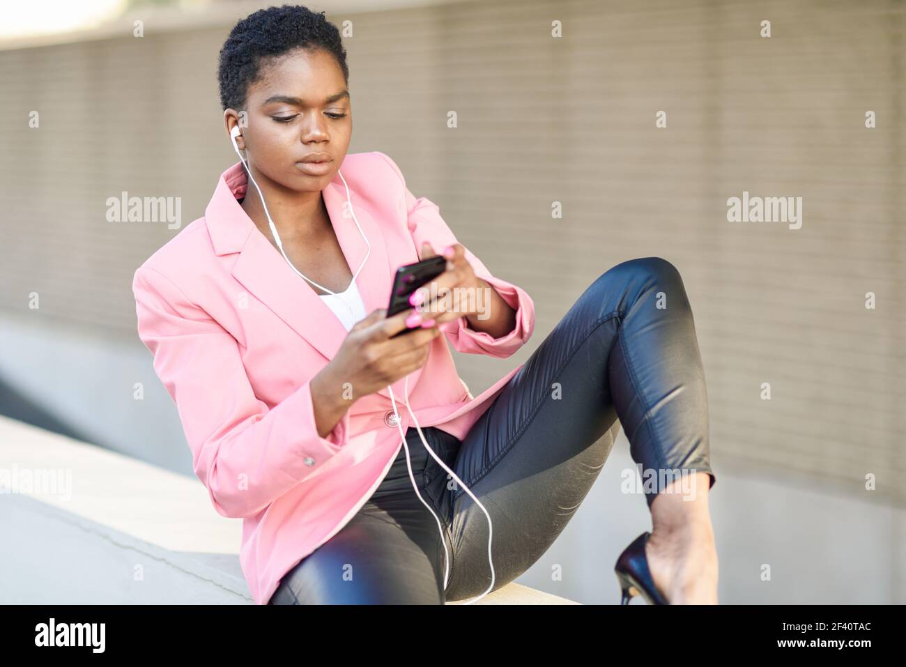 Donna d'affari nera seduta all'aperto utilizzando smartphone con auricolari. Donna afroamericana con giacca rosa. Donna d'affari nera seduta all'aperto utilizzando smartphone con auricolari Foto Stock