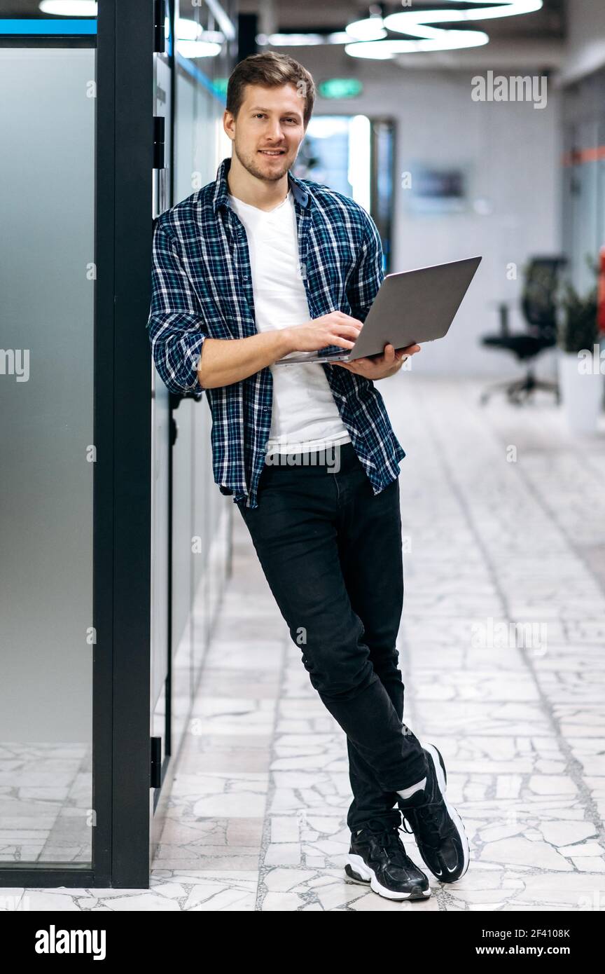 Caucasion freelance maschile è in piedi in ufficio moderno, guardando la fotocamera, sorridendo. Felice uomo d'affari giovane adulto in stile usa il laptop, lavorando su un nuovo progetto Foto Stock