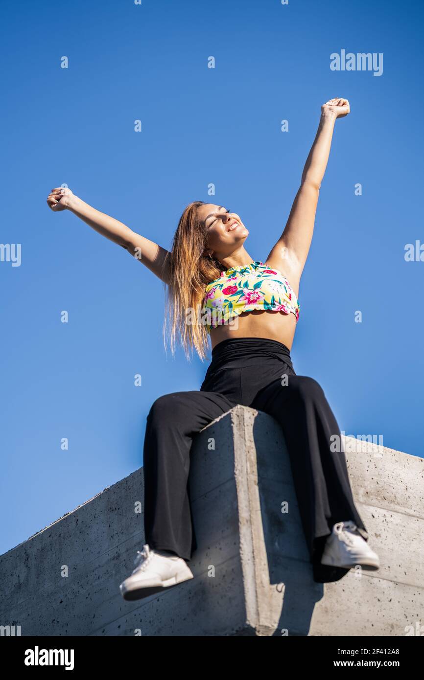 Giovane donna che apre le braccia contro il cielo blu seduta su muro urbano.. Giovane donna che apre le braccia contro il cielo blu Foto Stock