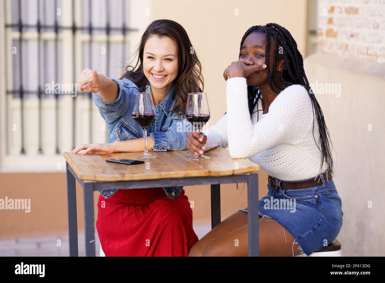 Due amici che bevono vino rosso seduti a un tavolo fuori da un bar. Donne multietniche.. Due donne che bevono vino rosso seduti a un tavolo fuori da un bar. Foto Stock