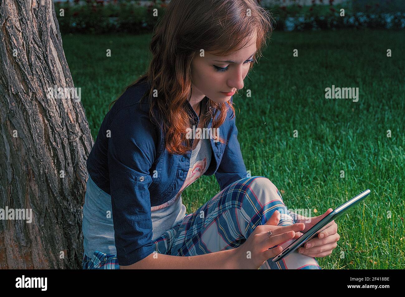 Ragazza che usa un computer tablet nel parco. Giovane donna rossa in calciato con un tablet pc nel parco che si appoggia contro un albero mentre scorre sul touchscreen. Ragazza che usa un computer tablet nel parco. Foto Stock