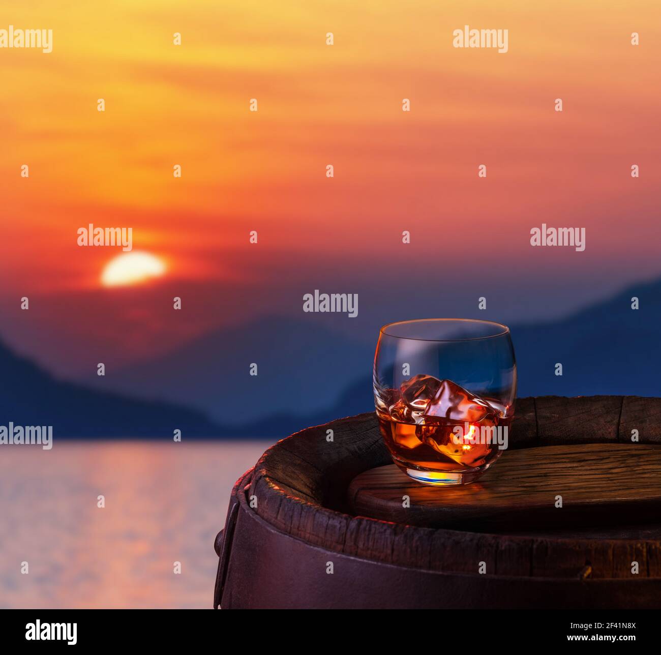 Bicchiere di rum scuro ghiacciato su barile di quercia. Rosso bel sole che scende sul mare sullo sfondo. Foto Stock