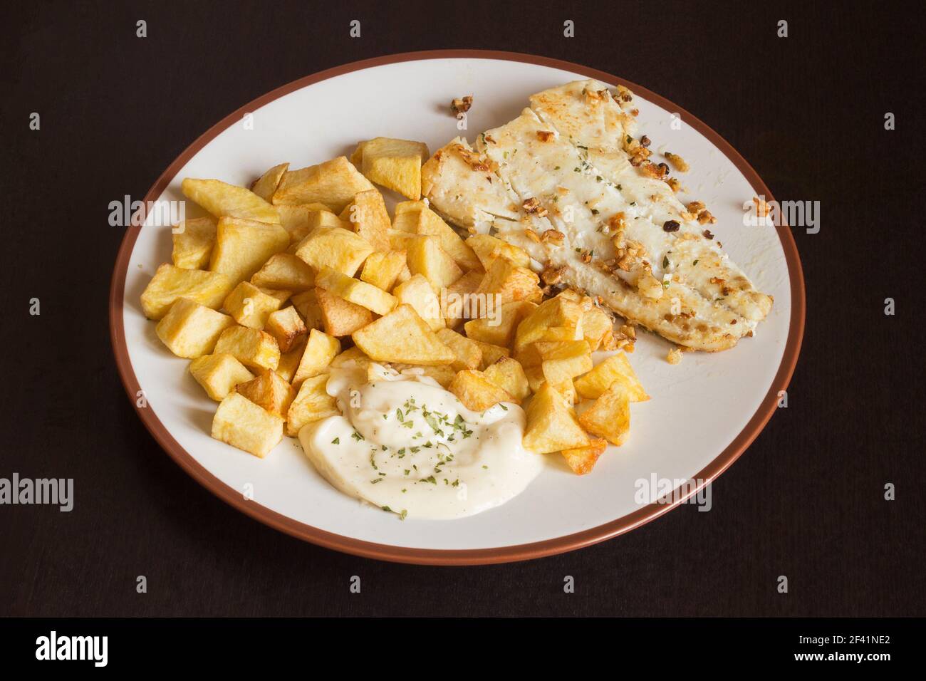 Condire con un filetto di nasello con aglio e prezzemolo accompagnato da salsa aioli e patate su un tavolo di legno scuro. Cibo e placcatura. Foto Stock