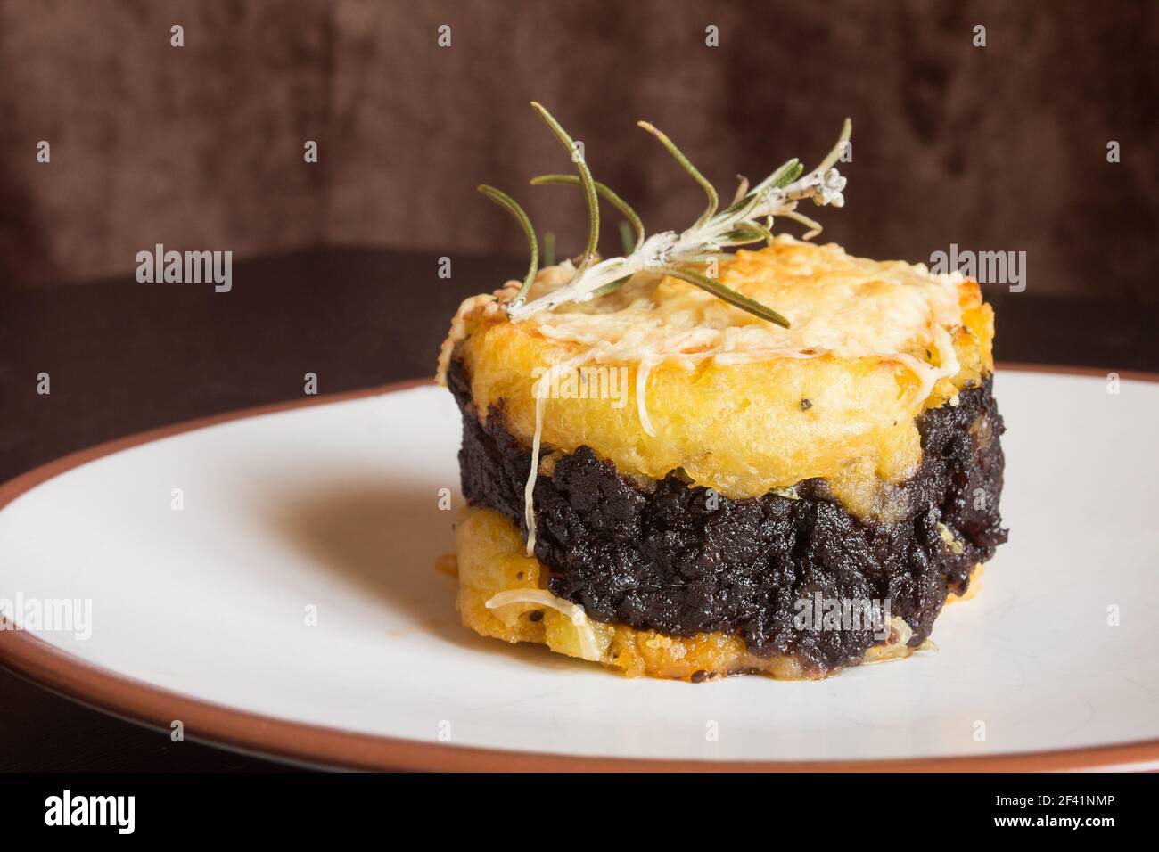 Primo piano di un sanguinoso pudding nero al cioccolato su un panino di patate cremoso con formaggio grattugiato e un fresco sprig di rosmarino sulla parte superiore di un tavolo di legno. Foto Stock