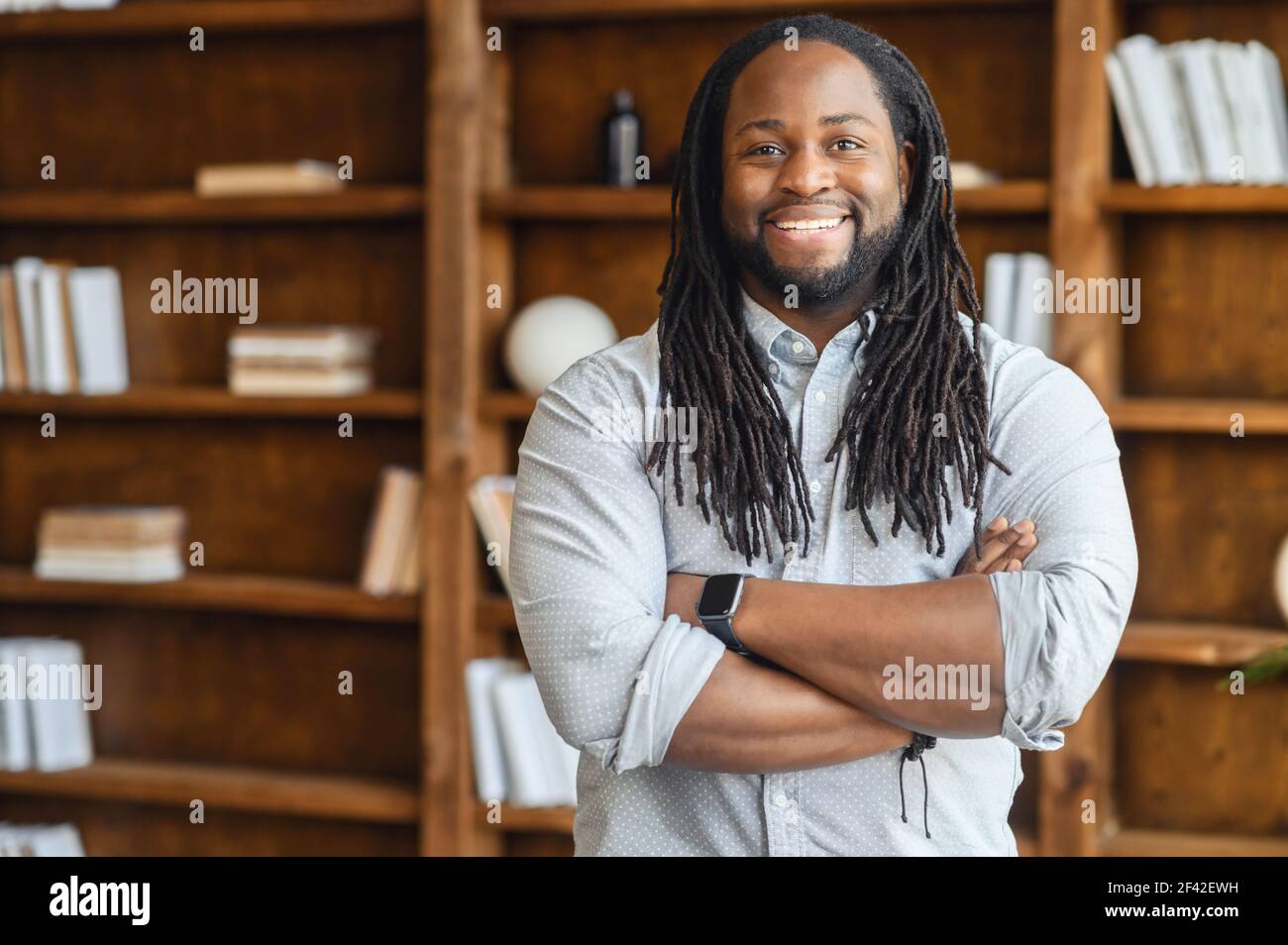 Ritratto di un giovane uomo africano sradicato con le spalle larghe e sorridente con i cavalletti con le braccia sul petto, guarda la macchina fotografica, sullo sfondo di scaffali con libri, freelance di successo Foto Stock
