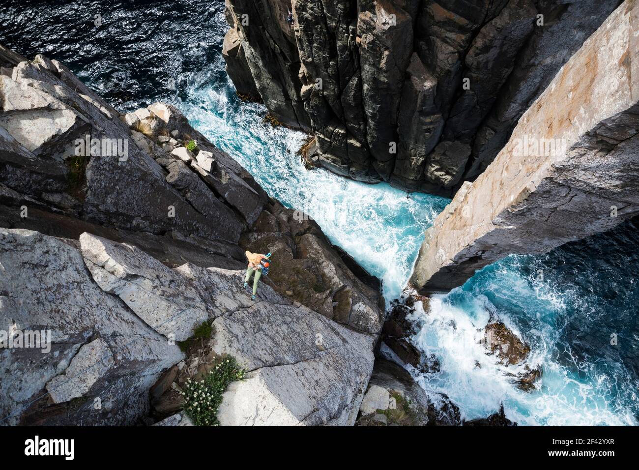 La rockclimber femminile guarda verso le onde che si infrangono mentre si precipita dal bordo di una scogliera di mare al fondo del Totem Pole, una colonna rocciosa che emerge dall'oceano Foto Stock