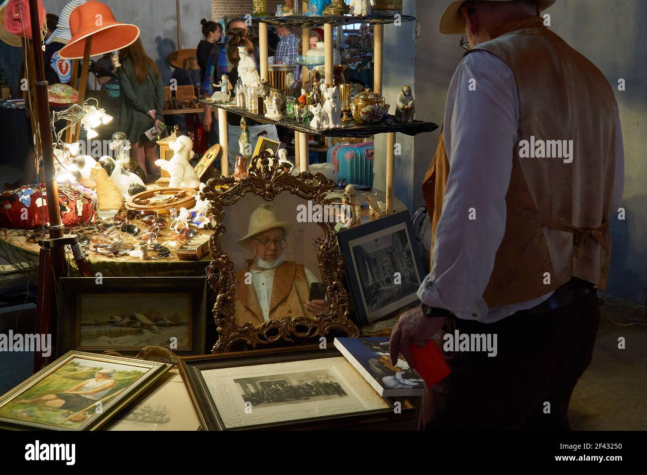 Riflesso di un uomo in un cappello largo-brimmed nello specchio su una bancarella al mercato.nel cortile del Museo di Mosca all'interno dell'anello del Giardino è il mercato delle pulci della città, un tesoro con stand di collezionisti e negozi di antiquariato che coesistono con i banchi di cittadini comuni che rappresentano l'antiquariato. Qui potete trovare quasi tutto - da porcellana antica, stoviglie e oggetti deco a gioielli, cartoline, badge e monete. Foto Stock