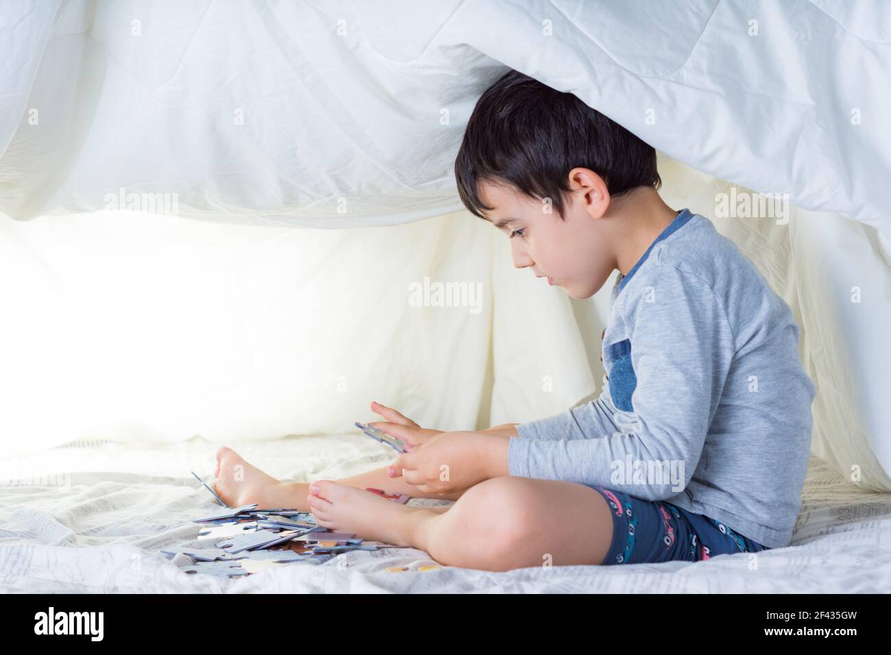 bambino che indossa pigiami dalle tonalità grigie in una tenda improvvisata con letto fogli che fanno un puzzle con sfondi bianchi e illuminazione soffusa Foto Stock