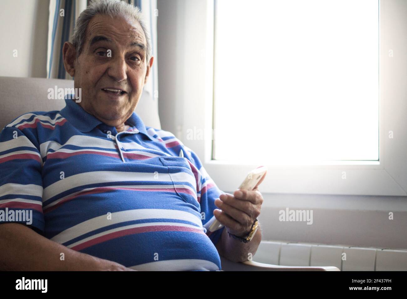 Persona anziana sorridente con smartphone seduto in una poltrona con una polo a righe accanto a una finestra molto luminosa Foto Stock