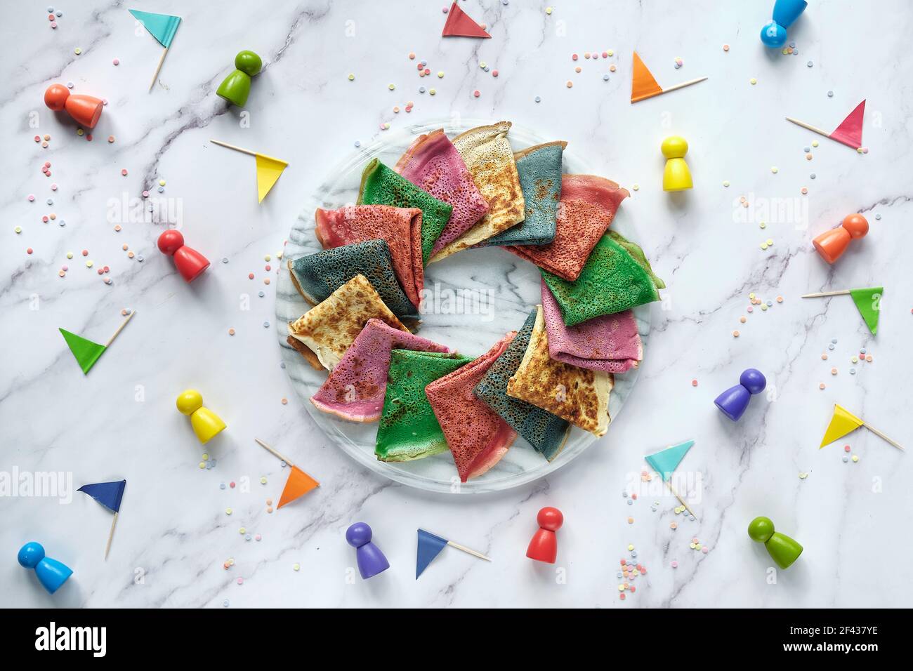 Rainbow party, tavolo di preparazione. Crepes vibranti su tavolo in marmo con figure in legno e bandiere di carta. Cibo creativo. Arcobaleno come simbolo di compassione Foto Stock