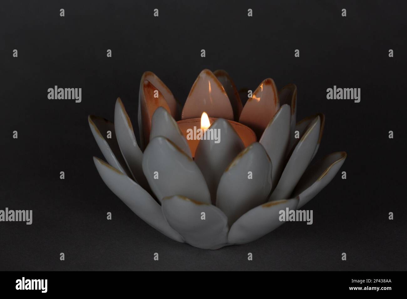 candela da tè illuminata con fiamma in un portacandele in ceramica di fiori di loto su uno sfondo grigio scuro con illuminazione drammatica e romantica e spazio per la copia Foto Stock
