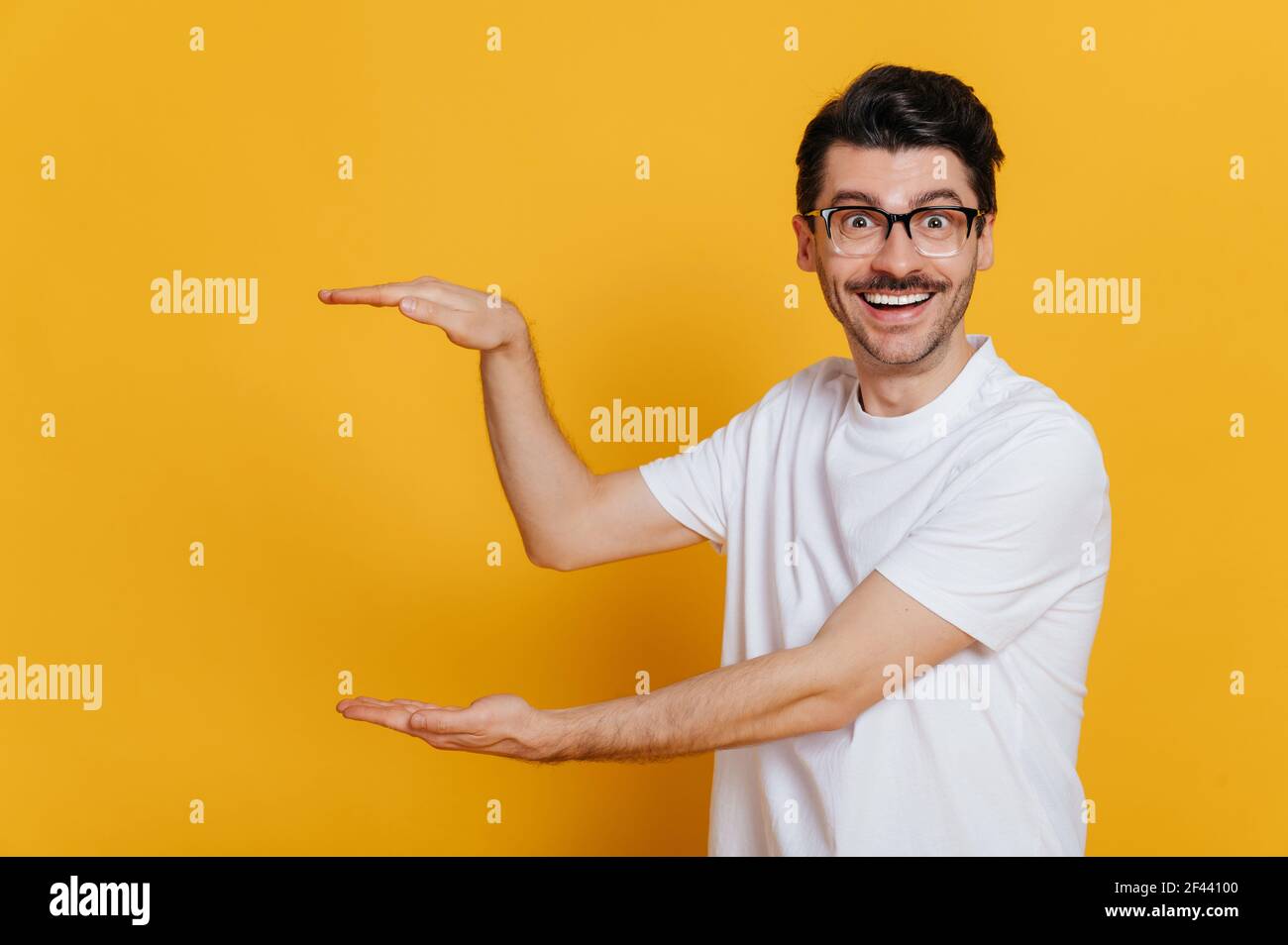 Scioccato eccitato felice caucasico unshaven giovane uomo adulto in bianco t-shirt e bicchieri mostrando con le mani ad uno spazio vuoto, in piedi contro isolato sfondo arancione, toothy sorridente Foto Stock