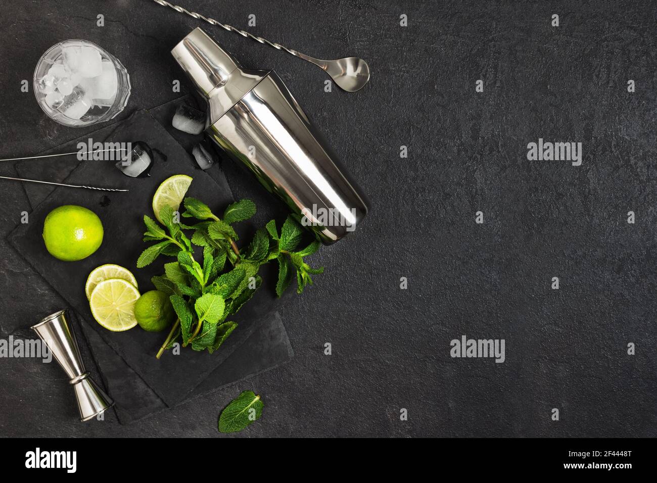Ingredienti e utensili da bar per preparare un cocktail mojito con cubetti di ghiaccio, menta fresca e lime su sfondo nero. Set di accessori per barre per mojito coc Foto Stock