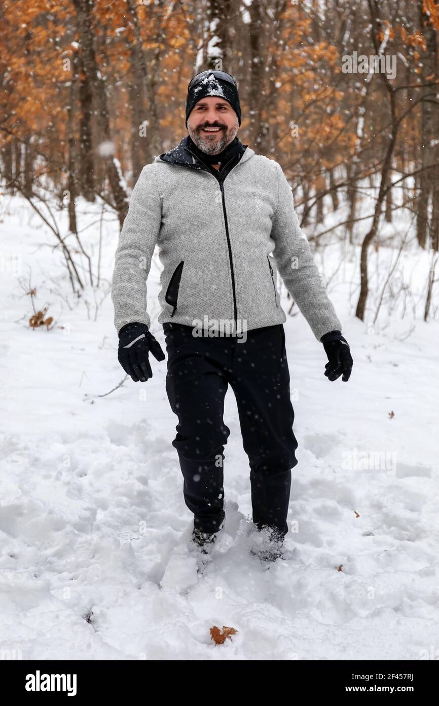 Un uomo bearded in abiti di montagna felpa grigia guanti e cappello nero immerso nella foresta innevata gioisce per i fiocchi di neve che cadono Foto stock Alamy
