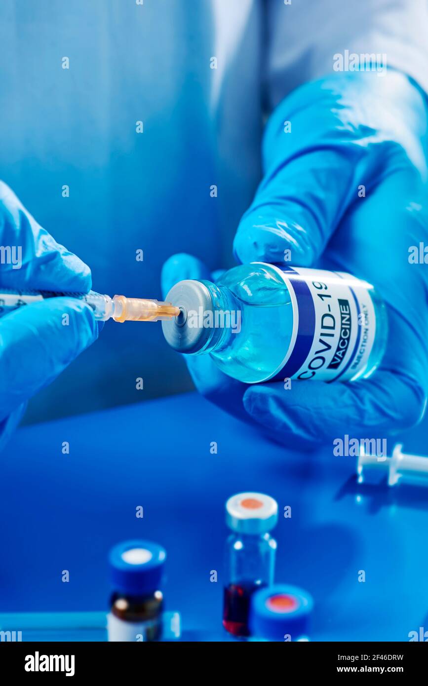 un operatore sanitario o di laboratorio, indossando guanti chirurgici blu, riempie una siringa con il vaccino covid-19 da un flaconcino simulato Foto Stock