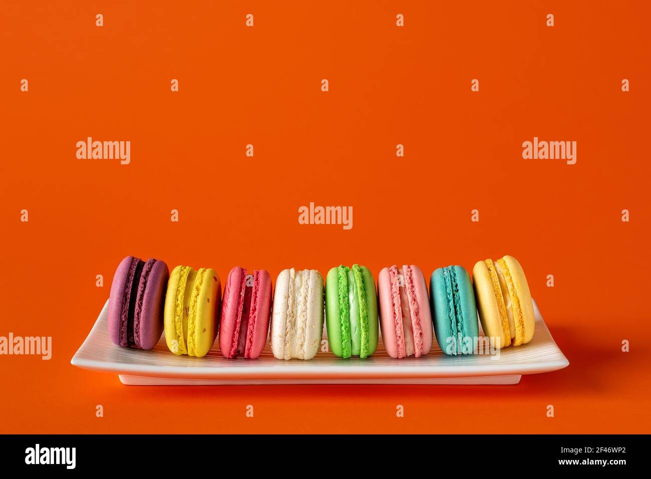 Macaroni multicolore in fila su una piastra rettangolare bianca su sfondo rosso brillante. Mockup di biglietto d'auguri con colorate torte macaron. Foto Stock