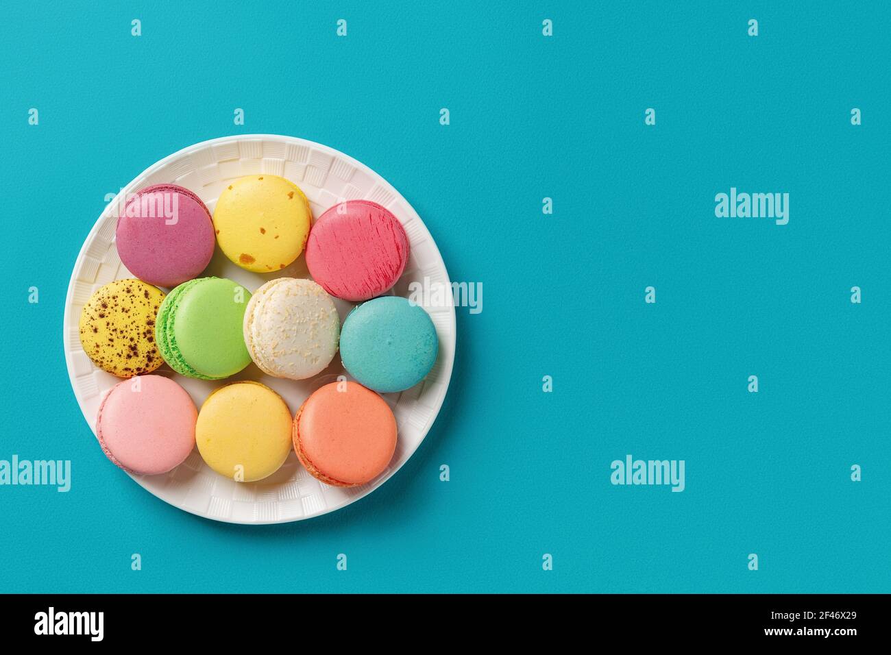 Macaroni colorati su una piastra rotonda bianca su uno sfondo turchese brillante. Mockup del biglietto d'auguri con dolci torte di macaron multicolore. Foto Stock
