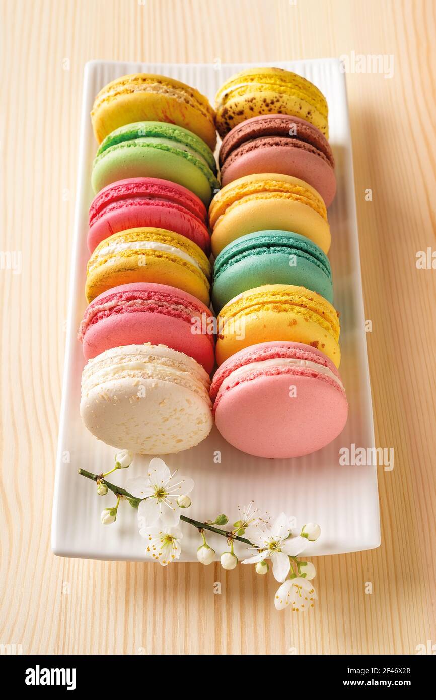 Set di amaretti multicolore e fiori di ciliegio primaverile su una piastra rettangolare bianca contro il tavolo in legno naturale. Gustosa meringa francese alle mandorle. Foto Stock