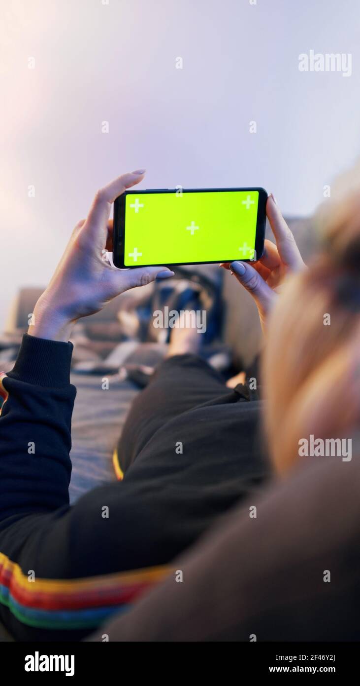 Giovane bionda che si rilassa sul divano e usa lo smartphone con schermo verde. Foto di alta qualità Foto Stock