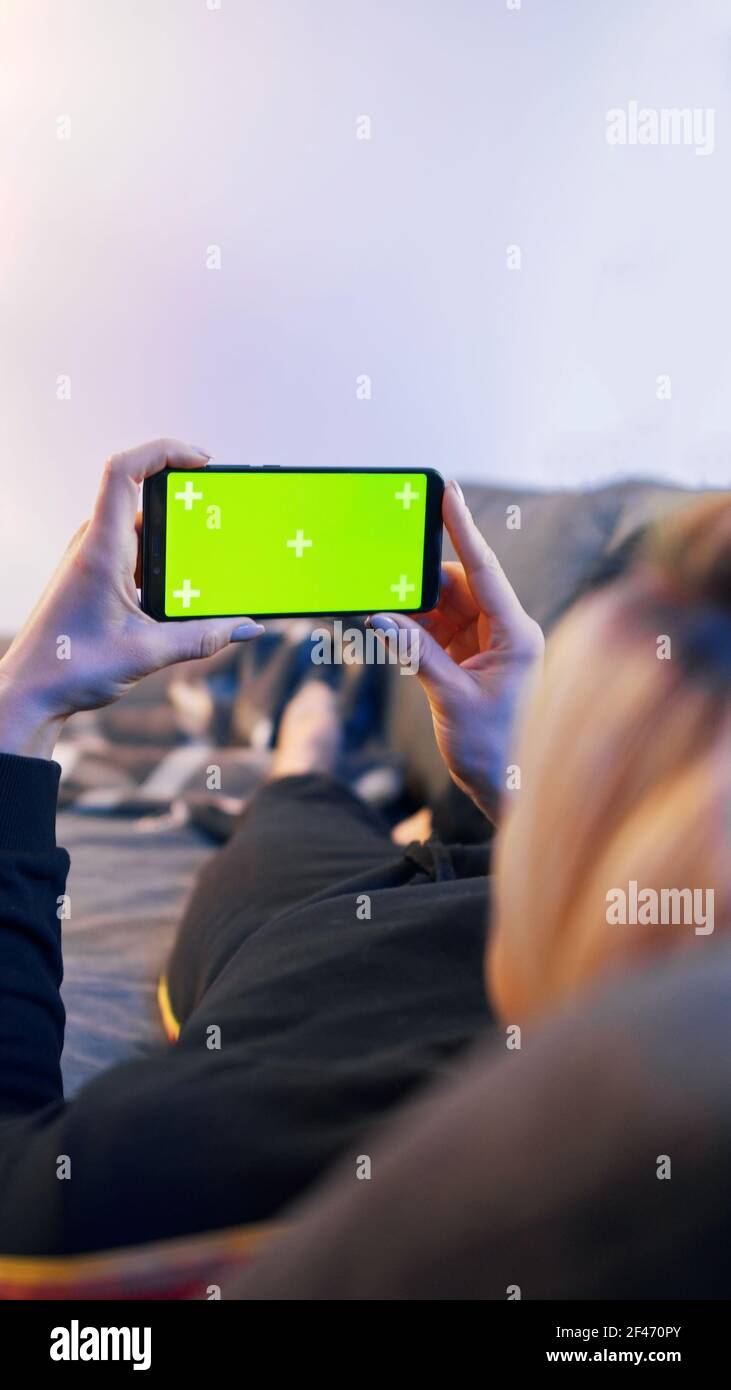 Giovane donna sdraiata su un divano e che utilizza uno smartphone con schermo orizzontale verde. Foto di alta qualità Foto Stock