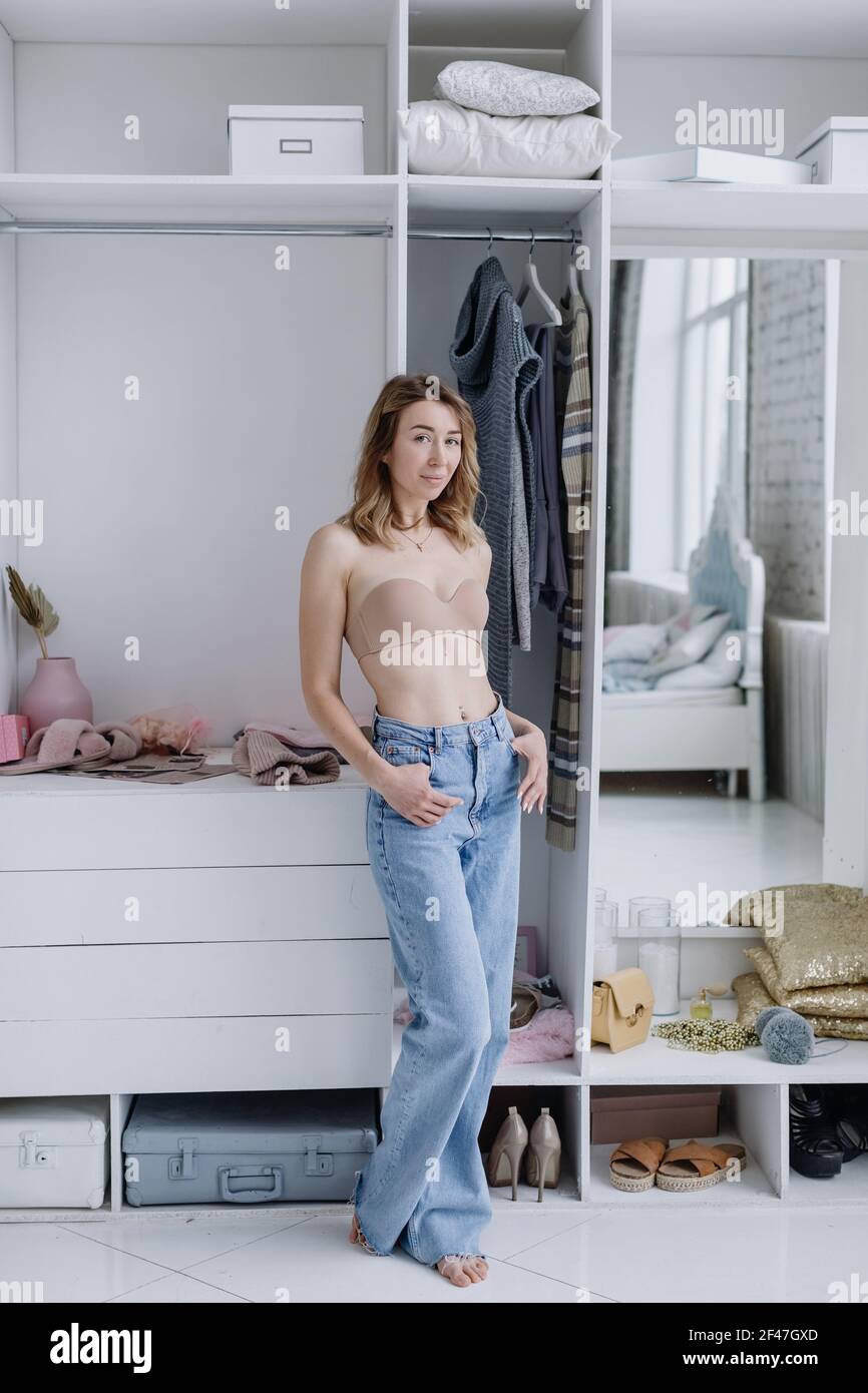 Bella giovane femmina in bodice beige, jeans in posa vicino armadio con vestiti in spogliatoio pensare che cosa indossare Foto Stock