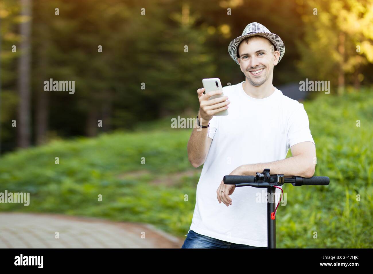 Giovane uomo in cappello usa il telefono cellulare, prenota online noleggio scooter elettrico in città. Trasporti innovativi Foto Stock