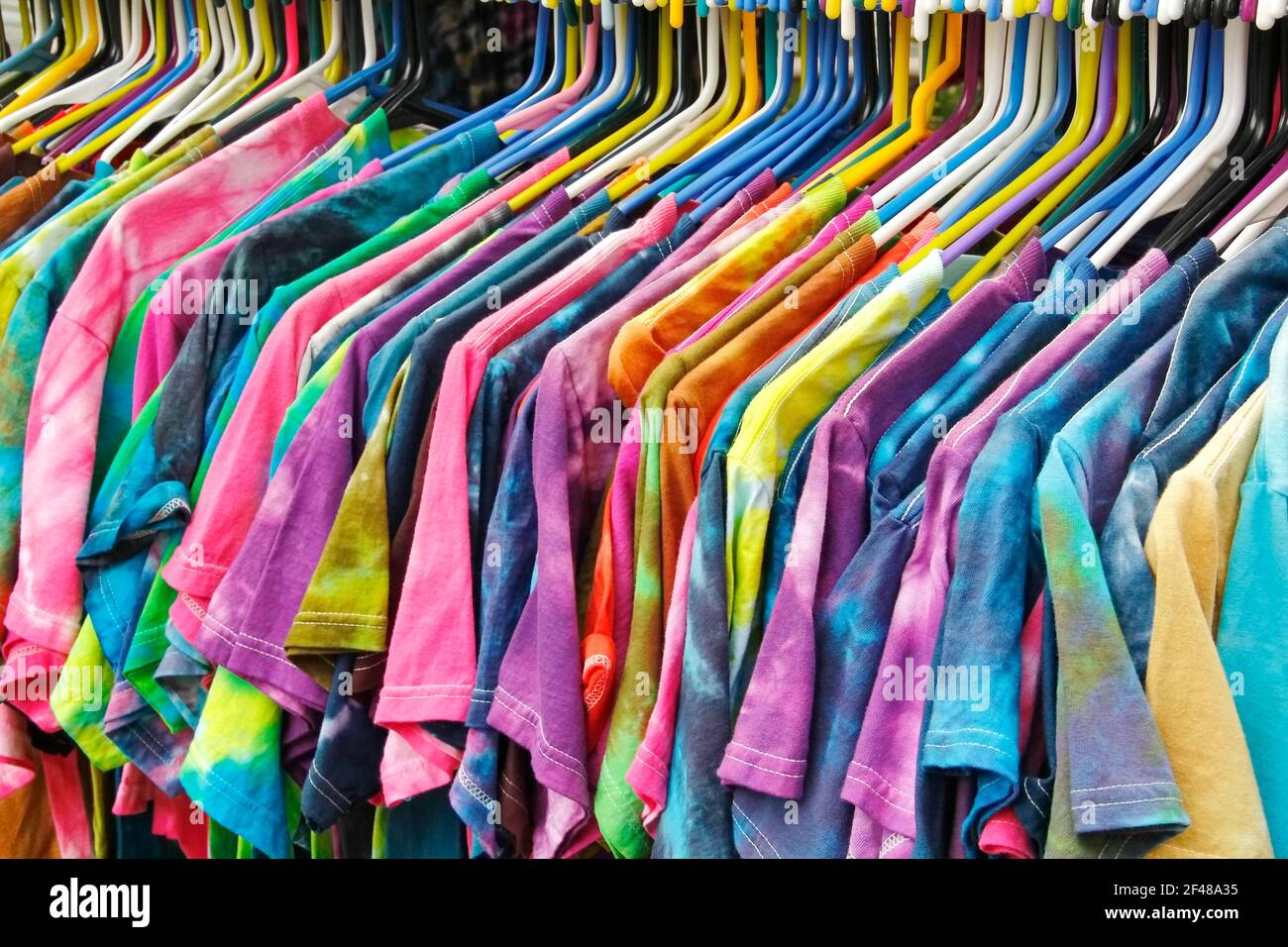 Vista laterale orizzontale di un rack di t-shirt tinte con cravatta di colore vibrante su staffe di plastica colorate allineate e appese in fila. Foto Stock