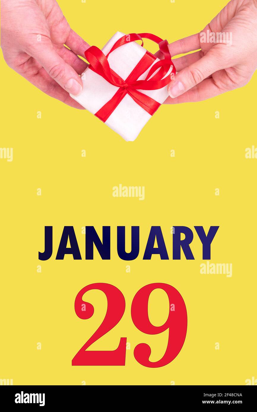 29 gennaio. Calendario verticale festivo con mani che tiene Bianco scatola regalo con nastro rosso e calendario Data 29 gennaio su illuminante Backgrou giallo Foto Stock