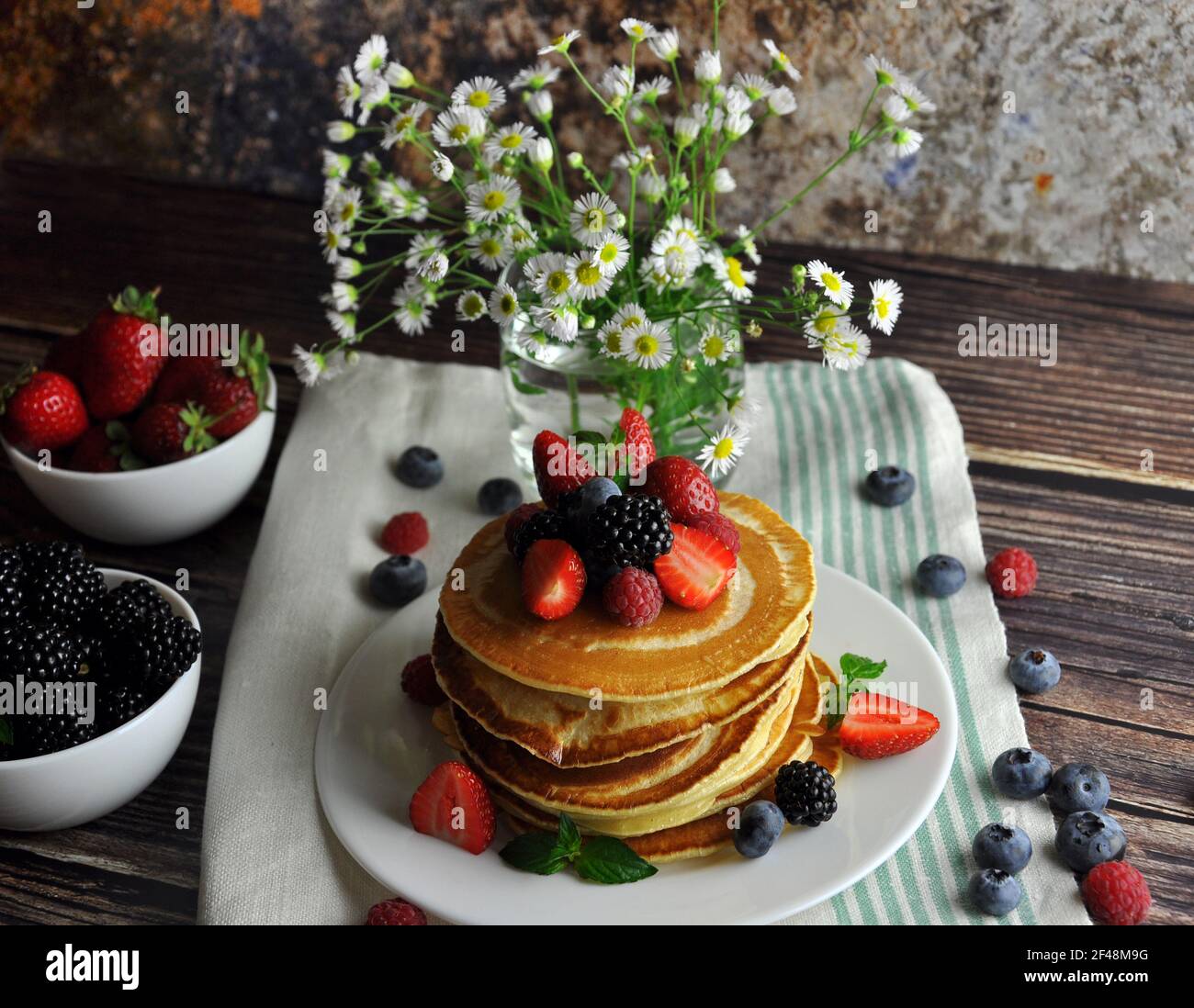 Pancake con frutti di bosco su un piatto bianco. Verticale. Vaso con fiori e frutta piccola. Foto Stock
