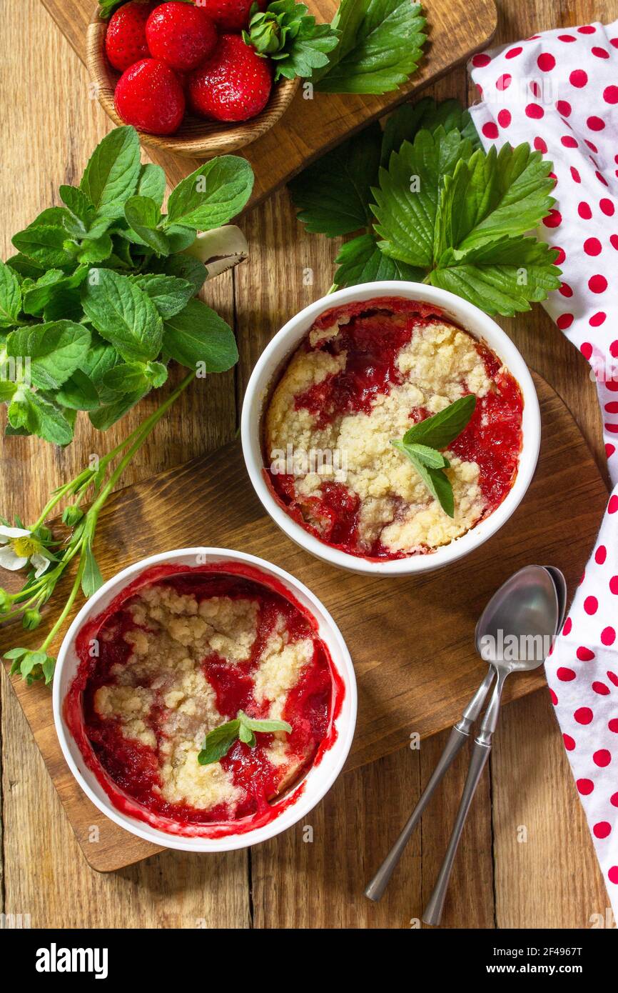 Dessert estivo cucina casalinga. Fragole sbriciolare in una teglia e fragole fresche su un tavolo rustico in legno. Vista dall'alto in piano. Foto Stock