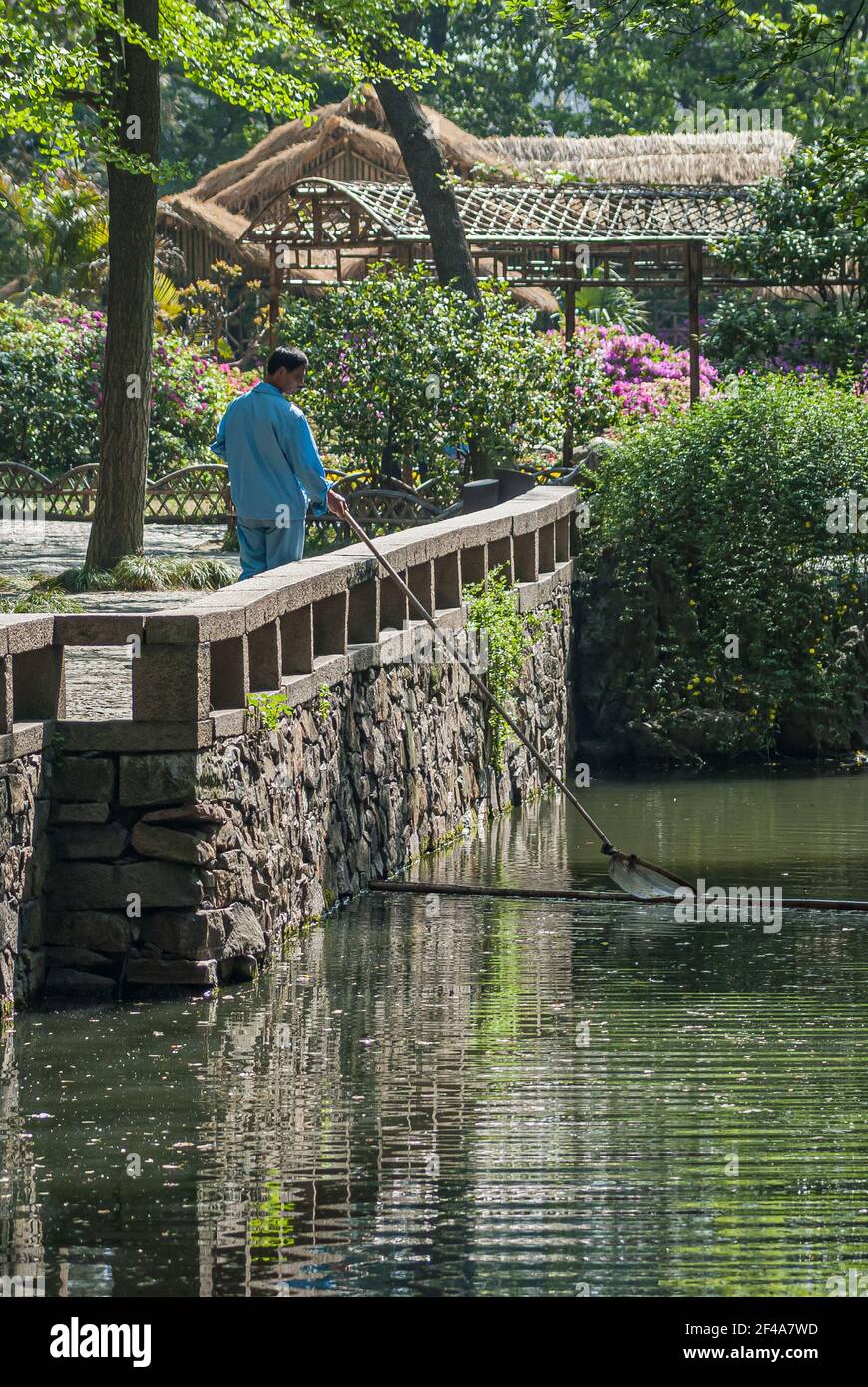 Suzhou, Cina - 3 maggio 2010: Giardino degli amministratori umili. Operaio in garb blu raccoglie i rifiuti di acqua verde insenatura in piedi sulla parete grigia di pietra con Foto Stock