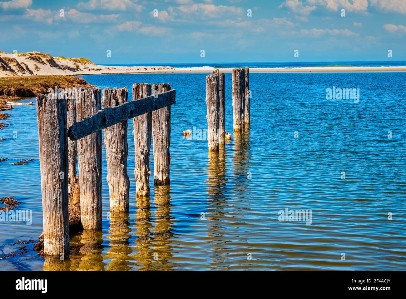 Resti decadenti di un molo in legno in acque blu con spiaggia sabbiosa e mare aperto oltre. Foto Stock