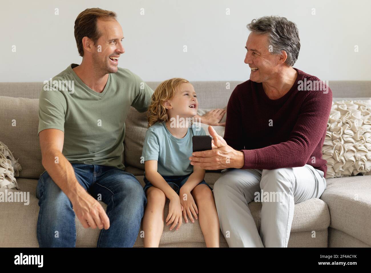 Nonno caucasico anziano sul divano che guarda lo smartphone con l'adulto figlio e nipote ridendo Foto Stock