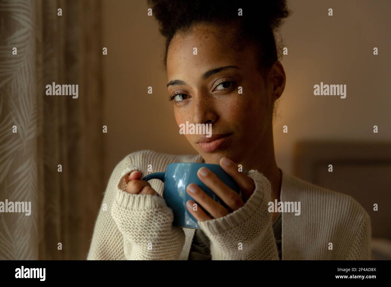 Donna di razza mista in piedi da una finestra e tazza da bere di caffè a casa Foto Stock