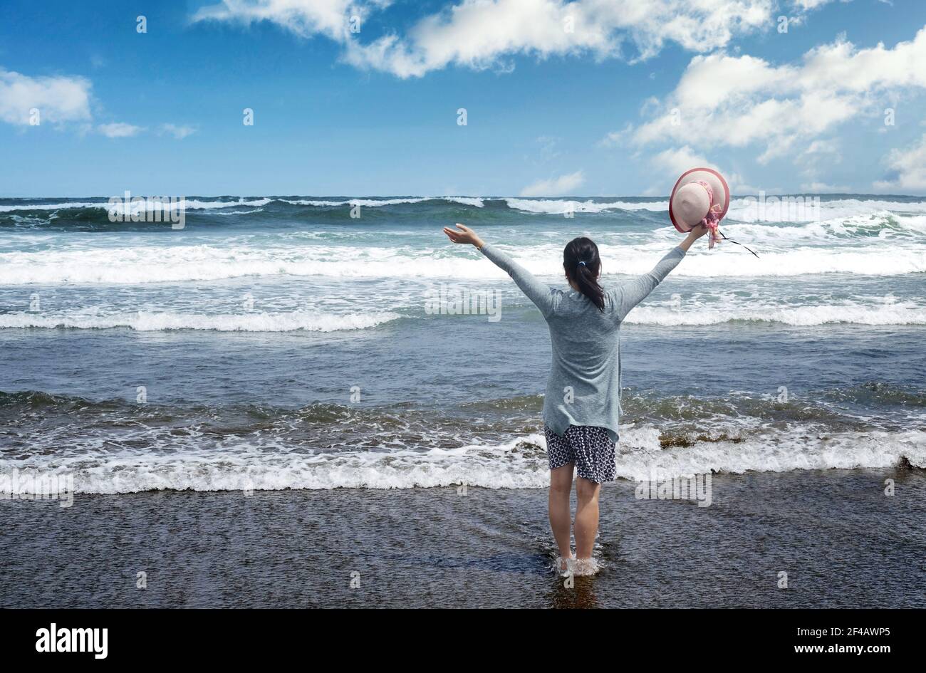 Donna di fronte oceano e cielo blu con le braccia in su mentre tenendo il cappello in espressione di pura gioia Foto Stock